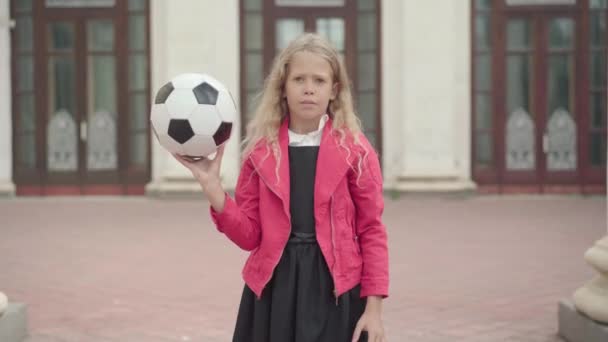 Jolie fille blanche blonde qui tourne la balle sur le doigt à l'extérieur. Portrait d'une écolière confiante et positive posant avec un équipement sportif de football ou de soccer. Sports féminins. — Video