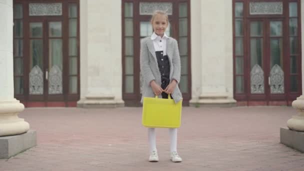 Широкий снимок счастливой девушки, стоящей на улице у входа в школу. Камера приближается к улыбающейся белой школьнице, позирующей после учебы. Концепция счастливого образования. — стоковое видео
