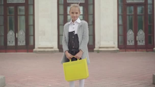 Egy mosolygó kaukázusi iskolás lány portréja, ahogy a kamerába néz, megfordul és iskolába sétál. Kamera követi boldog lány megy tanulni. Boldogság és oktatás. — Stock videók