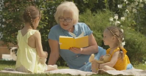 Retrato de mujer mayor caucásica en anteojos libro de lectura para niñas lindas al aire libre. Feliz abuela pasando el soleado día de verano con sus nietas. Sede del cine 4k ProRes. — Vídeos de Stock