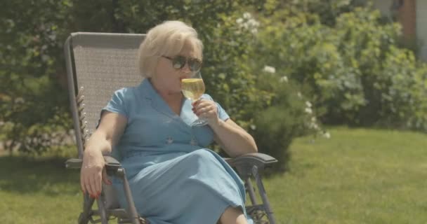 Pihentető, elegáns, idős nő ül a széken a szabadban és vizet iszik. Portré gondtalan kaukázusi hölgy napszemüvegben élvezi a napos nyári szabadnapot. Cinema 4k ProRes főhadiszállás. — Stock videók