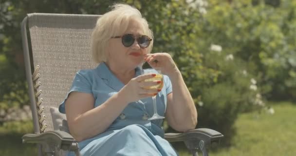 Happy senior retiree sitting in garden and drinking refreshing water. Portrait of positive carefree Caucasian woman in sunglasses enjoying sunny summer day outdoors. Cinema 4k ProRes HQ. — Stock Video