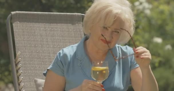 Retrato de una hermosa mujer mayor quitándose las gafas de sol y bebiendo agua de limón. Sonriente jubilado caucásico sentado en la silla del jardín y mirando a la cámara. Sede del cine 4k ProRes. — Vídeo de stock