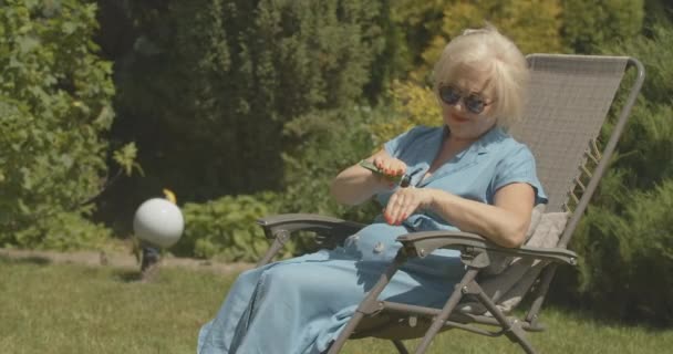 Despreocupado jubilado femenino sentado en silla de jardín y la aplicación de crema hidratante para las manos. Retrato de feliz señora caucásica relajada en gafas de sol tomando el sol al aire libre en un día soleado. Cine 4k proRes HQ. — Vídeos de Stock