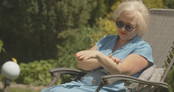 夏の庭でハンドクリームを適用するリラックスした女性の退職者。晴れた日に屋外で休んでサングラスでのんきな白人女性の肖像画。シネマ4k｜ProRes HQ. — ストック動画