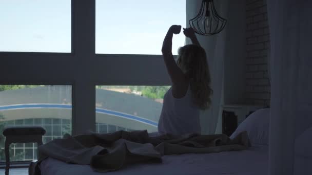 Hamile bir kadının silüeti sabahları yatak odasında geriniyor. Genç, beyaz, yakışıklı, evde uyanıp pencereden dışarı bakan bir adam. Hamilelik ve mutluluk. — Stok video