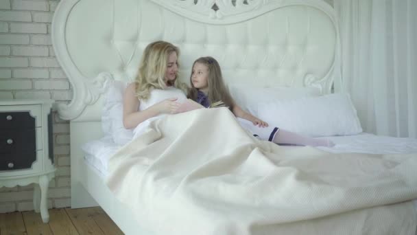 Szerokie ujęcie szczęśliwej ciężarnej matki leżącej w łóżku z córeczką i czytającej książkę. Portret młodej białej kobiety cieszącej się porankiem z uroczą dziewczyną w sypialni. Koncepcja rodziny. — Wideo stockowe