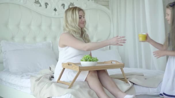 Bruna ragazza portando succo d'arancia per la madre incinta seduta sul letto con l'uva. Ritratto di donna incinta caucasica felice che riposa a casa con la figlia premurosa. Concetto di gravidanza. — Video Stock