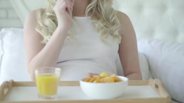 Nierozpoznana biała kobieta jedząca niezdrowe chipsy ziemniaczane i głaskająca brzuchem ciężarnej. Beztroska przyszła matka, siedząca rano na łóżku i jedząca śniadanie. Koncepcja uszkodzenia płodu. — Wideo stockowe