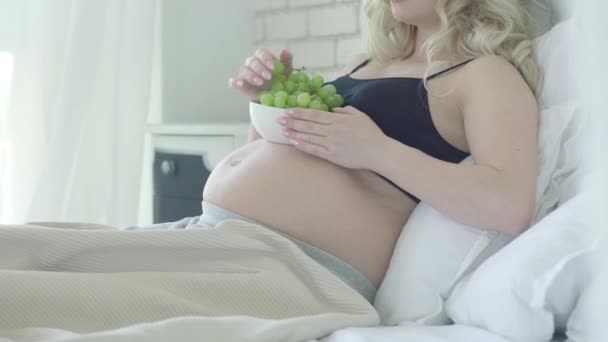 Femme enceinte méconnaissable couchée sur le lit et mangeant des raisins sains. Dame caucasienne détendue appréciant la nourriture saine à la maison dans la chambre. Concept de grossesse heureuse. — Video