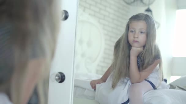 Triste petite fille regardant dans le miroir et soupirant. Reflet d'un enfant brune caucasien bouleversé avec les yeux bruns assis sur le lit à la maison et pensant. Enfant malheureux seul à l'intérieur. — Video