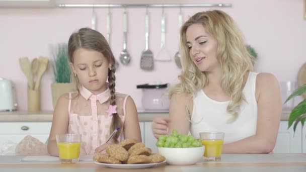 Mooie jonge moeder helpt dochter met huiswerk in de ochtend. Portret van een gelukkige blanke ouder en kind zittend aan tafel in de keuken en pratend. — Stockvideo