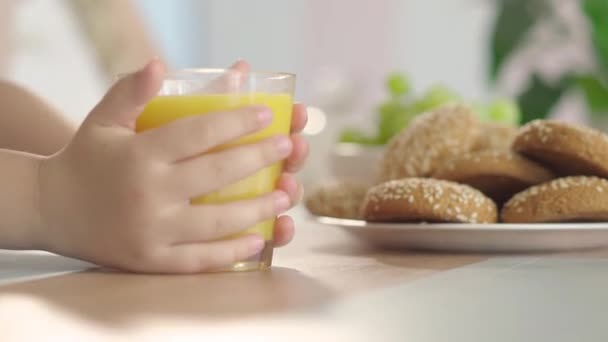 Pequeños niños mano tomando un vaso de jugo de naranja de la mesa. Niño caucásico irreconocible bebiendo bebida saludable como mujer borrosa sentada en el fondo. — Vídeo de stock