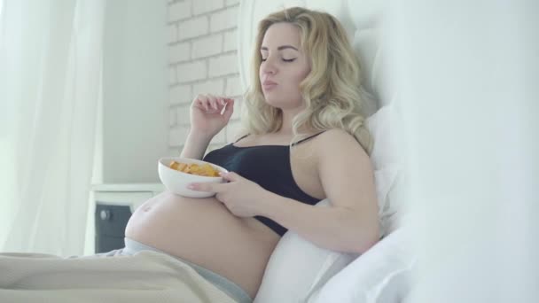 Onvoorzichtige zwangere vrouw die geroosterde chips kauwt. Portret van zorgeloze blanke verwachting liggend op bed en het eten van schadelijk ongezond voedsel. Voeding tijdens de zwangerschap. — Stockvideo