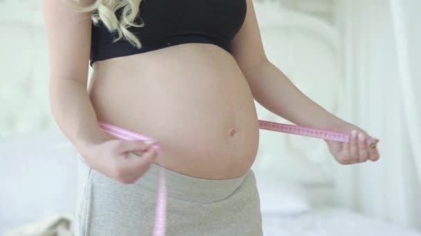 Femme enceinte mesurant le ventre et souriant à la caméra. Heureux souriant Caucasien attendant bébé. Belle dame blonde debout dans la chambre à la maison. Jolie grossesse et maternité. — Video