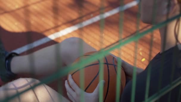 Bovenaanzicht van onherkenbare sportman zittend met bal in het zonlicht op het buitenveld. Jonge blanke man rust na de sportwedstrijd op zonnige zomerdag buiten. Gezonde levensstijl. — Stockvideo