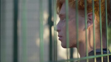 Güneş ışığında tel örgülerin arkasındaki kızıl saçlı genç adamın yakın çekimi. Güneşli bir yaz gününde açık havada beyaz bir adamın portresi..