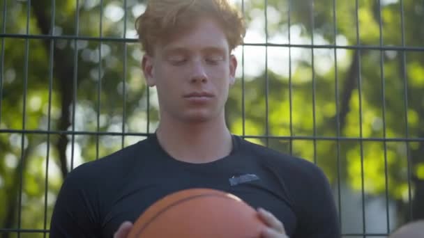 Retrato de jogador de basquete ruiva pensativo segurando bola laranja e olhando para longe na luz do sol. Confiante chateado desportista caucasiano inclinando-se contra cerca de malha em ensolarado tribunal ao ar livre e pensando. — Vídeo de Stock