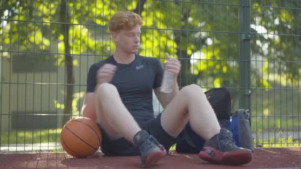 Vermoeide sportman die verfrissend water drinkt na het trainen buiten. Portret van een jonge roodharige blanke man die na basketbal op het veld zit. Sportieve levensstijl. — Stockvideo
