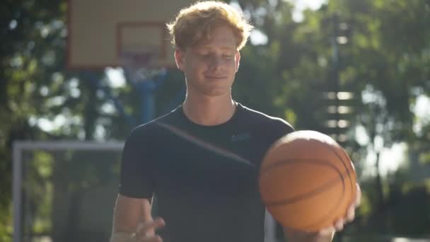 Vreugdevolle jonge sporter poseren met bal in het zonlicht op het buitenveld. Portret van een glimlachende roodharige blanke man die op de achtergrond van de basketbalring staat en naar de camera kijkt. — Stockvideo