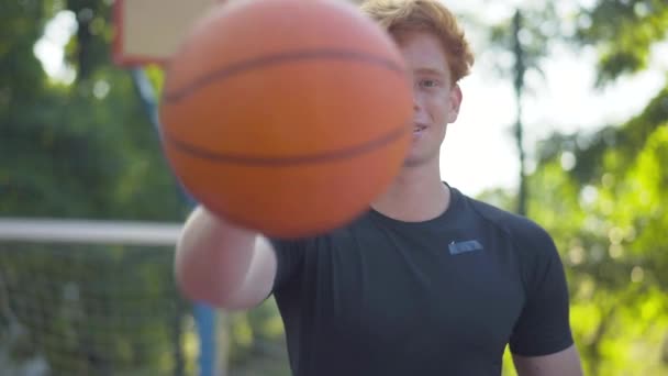 Zelfverzekerde blanke basketbalspeler die de bal naar de camera uitrekt en glimlacht. Portret van een jonge vrolijke man die poseert op het zonnige buitenveld op zomerdag. Sport en workout concept. — Stockvideo