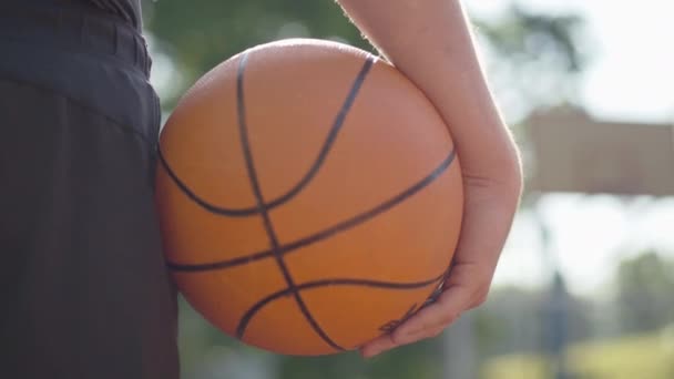 Close-up de bola de basquete na mão masculina à luz do sol. Desportista caucasiano irreconhecível em pé em raios de sol ao ar livre com equipamento desportivo. Confiança e treino. — Vídeo de Stock