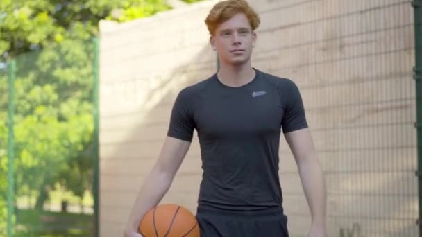 Knappe zelfverzekerde basketbalspeler die met bal naar camera loopt en vertrekt. Portret van een jonge blanke sportman die op zonnige zomerdag buiten slentert. — Stockvideo