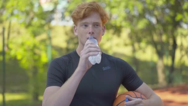 Portré izzadó sportolóról, aki frissítő vizet iszik az üvegből és a kamerába néz. Fiatal vörös hajú férfi pózol labdával a szabadban a sportos játék után. Aktív életmód koncepció. — Stock videók