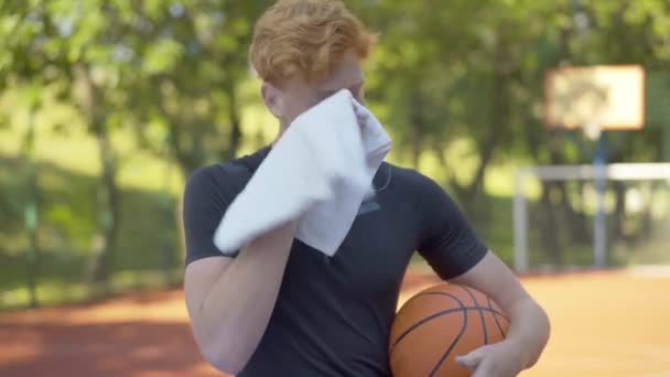 Zwevende sportman veegt gezicht met witte handdoek en zuchten. Portret van de jonge roodharige blanke man op het buitenveld met basketbal. Sport en mannelijkheid. — Stockvideo