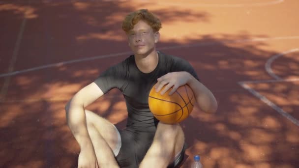 屋外のコートでボールと座って日光を見上げ疲れ汗をかく赤毛のスポーツマン。バスケットボール大会の後に屋外で休んで疲れた若い白人男性の肖像画. — ストック動画