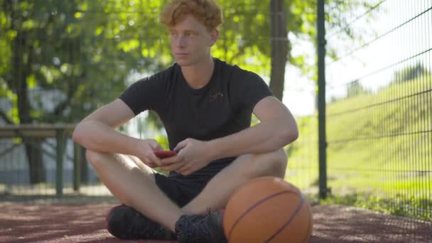 Pemain bola basket berambut merah yang bijaksana duduk di lapangan terbuka dan menggunakan smartphone. Potret santai pria Kaukasia beristirahat setelah pelatihan luar ruangan surfing Internet. — Stok Video