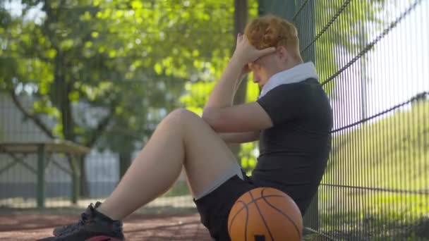 Zijaanzicht van uitgeputte basketbalspeler zittend aan mesh hek met hoofd in handen. Portret van blanke man losser na sport spel op het buitenveld in zonlicht. — Stockvideo