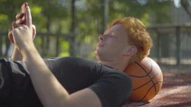 Kaygısız basketbolcunun güneş ışığında topun üzerine uzanması ve akıllı telefon kullanması. İyi gevşetilmiş Kafkasyalı kızıl saçlı adam idmandan sonra internetten mesajlaşıyor..