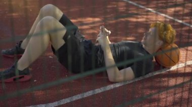 Tel örgülerin arkasında top üzerinde uzanan ve akıllı telefondan mesajlaşan rahat bir adamın geniş açılı görüntüsü. Sosyal medyayı açık alanda dinlenmek için kullanan genç, pozitif kızıl saçlı beyaz sporcu..