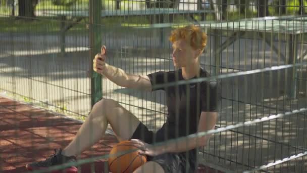 Gelukkig ontspannen sporter nemen selfie en glimlachen als zittend op het buitenveld op mesh hek. Positieve roodharige blanke man met smartphone op zonnige zomerdag na het sporten buiten. — Stockvideo