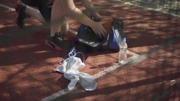 Deportista caucásico irreconocible que empaqueta la pelota, la camisa y los auriculares en una bolsa deportiva. Joven hombre caucásico reuniendo equipo después del partido de baloncesto en la cancha al aire libre. Deporte y estilo de vida. — Vídeo de stock