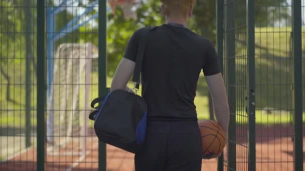 Achteraanzicht van zelfverzekerde sporter die de buitenbaan betreedt met sporttas en bal. Jonge Kaukasische basketbalspeler wandelend voor training op zonnige zomerdag buiten. Sport en workout. — Stockvideo