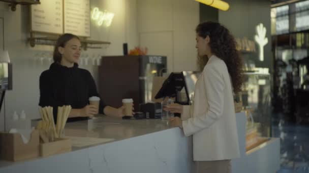 Mulher branca sorridente servindo café para ir para a jovem mulher de negócios bonita no café. Mulher positiva barman caucasiano vendendo takeaway bebida quente no restaurante. — Vídeo de Stock