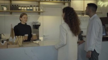 Pozitif kafe işçisinin portresi. Tezgahın üstünde müşterilere kahve ve kağıt torba verip gülümsüyor. Kafkasyalı kadın ve erkek yemek ve içecekle ayrılıyorlar. Hizmet ve restoran işi.