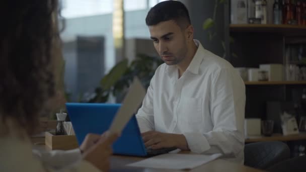 Portrait d'un homme d'affaires confiant du Moyen-Orient utilisant un ordinateur portable et parlant avec une femme d'affaires méconnaissable dans un café. Un jeune homme concentré discute de la coopération au restaurant le midi. — Video