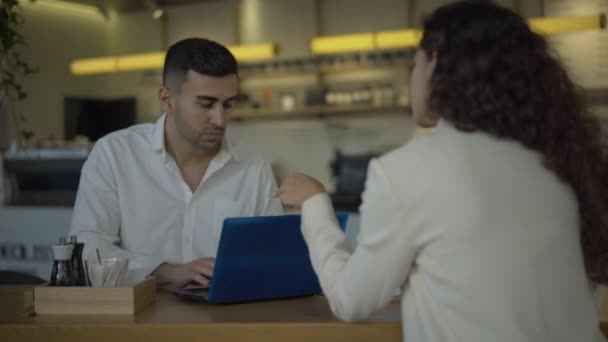 Knappe jonge zakenman uit het Midden-Oosten die in een café zit met een blanke zakenvrouw en laptop gebruikt. Portret van een succesvolle man die praat met een vrouw en surft op internet. Zakelijk lunchconcept. — Stockvideo