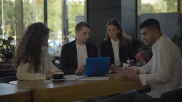 Vier multi-etnische jongeren die zakenlunchen in een café. Portret van drukke zelfverzekerde Kaukasische en Midden-Oosterse mannen en vrouwen die surfen op internet en discussiëren over teamwork. Samenwerkingsconcept. — Stockvideo