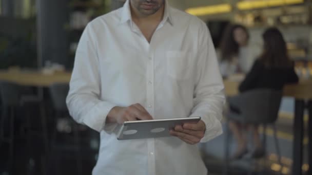 无法辨认的中东商人穿着白衬衫，用平板电脑在网上发短信。自信忙碌的男性创业者在网上冲浪，背后有模糊的咖啡馆或餐馆. — 图库视频影像