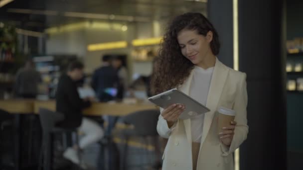 Retrato de sucesso jovem empresária posando com tablet e xícara de café. Sorrindo bela mulher caucasiana beber takeaway bebida quente com pessoas desfocadas no café no fundo. — Vídeo de Stock