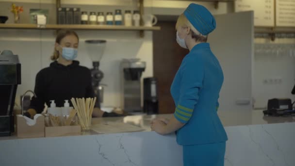 Sebevědomá žena v uniformě letušky si objednává jídlo v kavárně a odchází. Mladý kavkazský barman v Covid-19 s obličejovou maskou předává papírovou tašku a kávu. Koronavirový životní styl. — Stock video