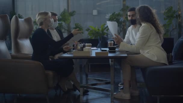 Gros plan de quatre hommes d'affaires portant des masques Covid-19 assis à la table dans un café et parlant. Chers collègues, discuter de l'accord sur la pause café pendant le confinement en cas de pandémie de coronavirus. — Video