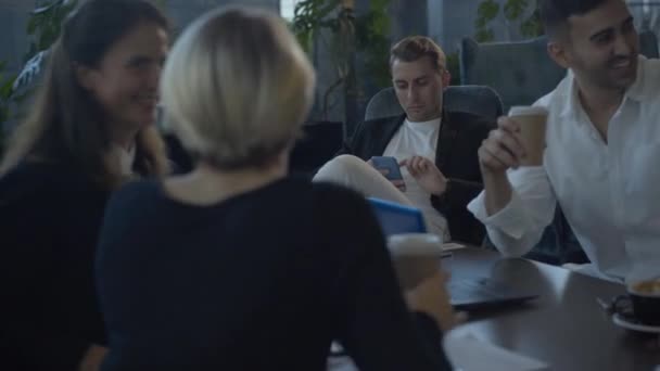 Bored Caucasian man using smartphone while cheerful colleagues talking and laughing at the table in cafe. Portrait of handsome successful businessman uninterested in conversations with coworkers. — Stock Video