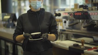 Covid-19 yüz maskeli, elinde kahve ya da çay fincanı tutan koruyucu eldivenli, tanınmayan beyaz bir kadın. Coronavirus salgınında çalışan profesyonel bir barmen..