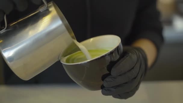 Αγνώριστος μπάρμαν προσθέτοντας γάλα στο πράσινο τσάι matcha. Κοντινό πλάνο των χεριών σε προστατευτικά μαύρα γάντια ανάμειξη υγιεινό ζεστό ρόφημα στο μπαρ στο εστιατόριο ή καφέ. — Αρχείο Βίντεο