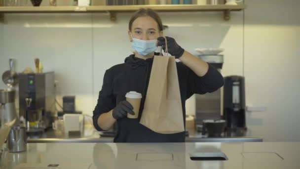 Wanita kaukasia yang percaya diri dengan topeng wajah Covid-19 membentangkan cangkir kopi dan kantong kertas di depan kamera. Potret karyawan profesional yang melayani orang di kafe atau restoran selama pandemi coronavirus. — Stok Video